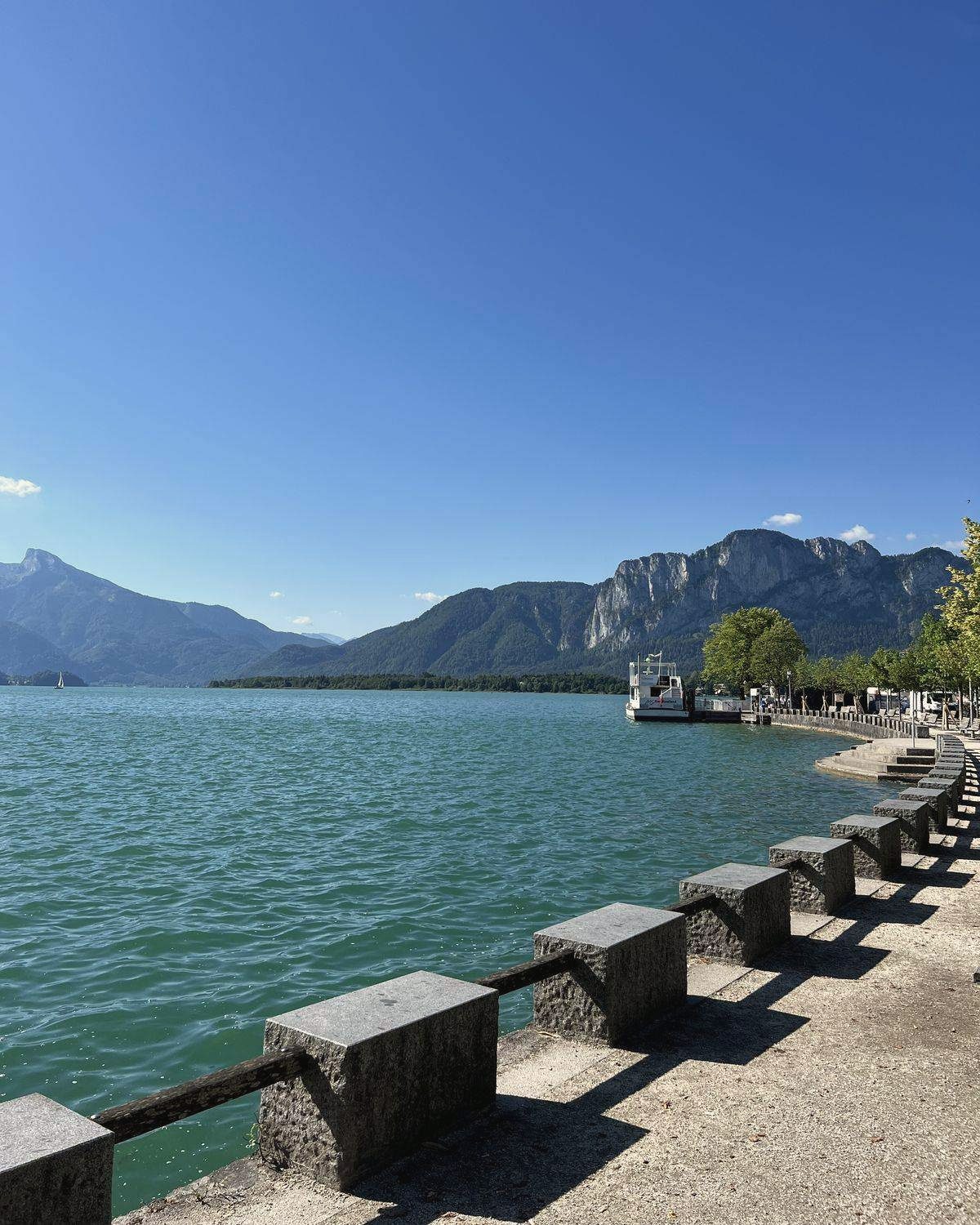 Türkisfarbener See mit Uferpromenade und Bergen im Hintergrund